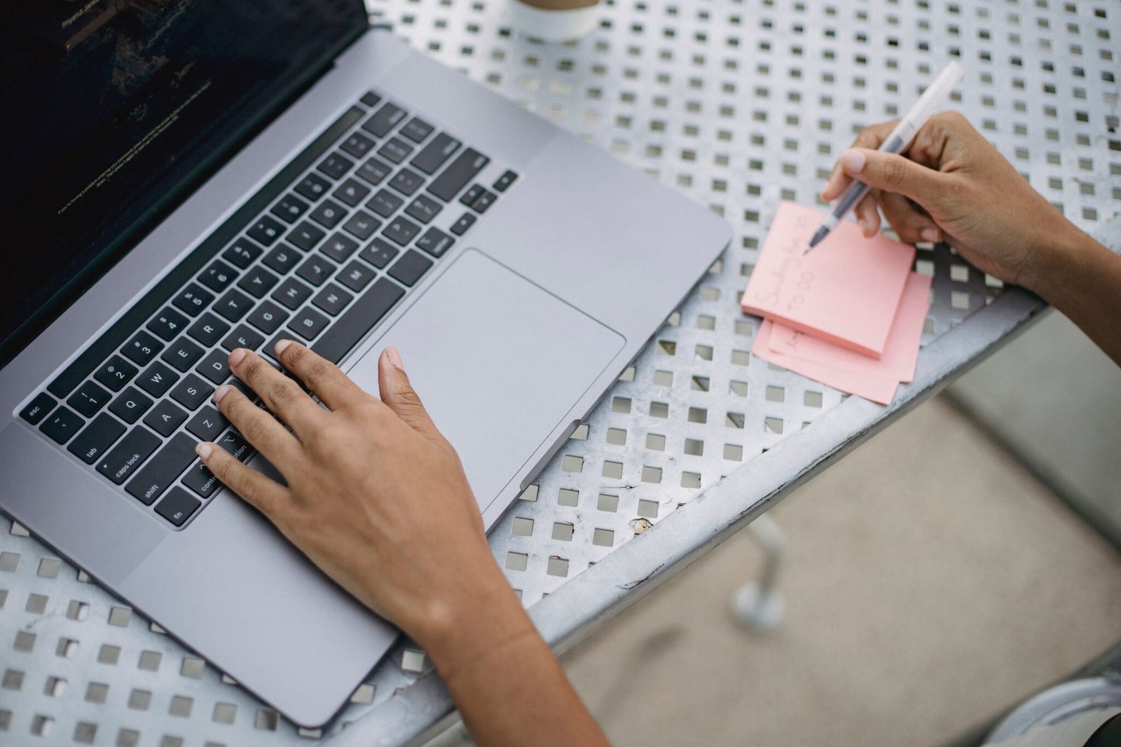 Close-up of a person using a laptop and writing notes at an outdoor workspace.